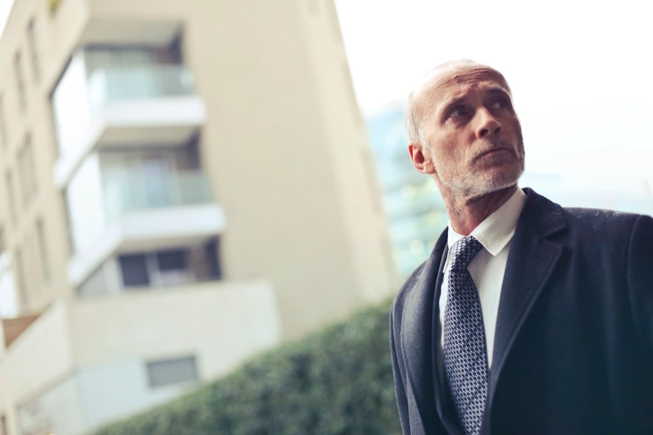 A serious businessman in a formal suit and tie stands outdoors in Milan, looking upwards thoughtfully.